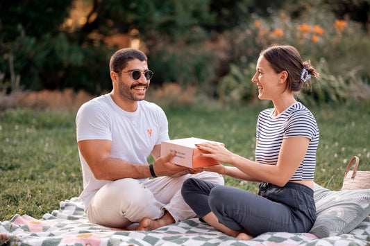 Man and woman sitting on a blanket in a park, exchanging a gift box.